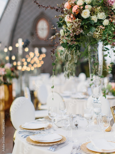 Wedding table setting in gold and white. On the table is a white tablecloth, dishes, cutlery, and a bouquet of flowers in a tall glass vase. In the background is a brown tower with an oval window.