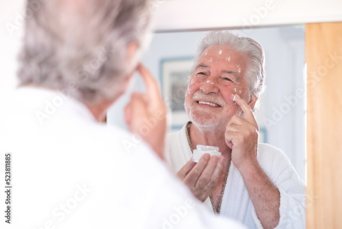 Skin care. Handsome senior man applying cream at his face and looking at himself with smile while standing in front of the mirror