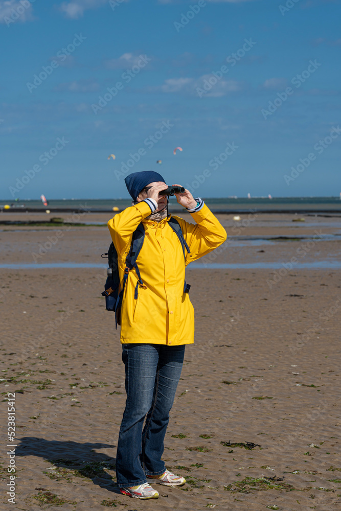 Langrune-Sur-Mer, France - 08 05 2022: Portrait of a woman wearing a yellow raincoat, a blue head cover, a sand scarf, sunglasses, binoculars and a backpack at the beach