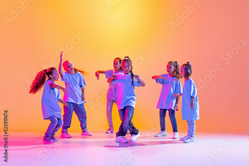 Group of children, little girls in sportive casual style clothes dancing in choreography class isolated on orange background in purple neon light.