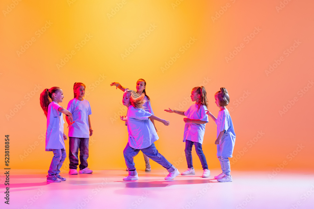 Portrait of cheerful, active little girls, happy kids dancing isolated on orange background in neon light. Concept of music, fashion, art, childhood, hobby