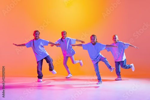 Portrait of cheerful  active little girls  happy kids dancing isolated on orange background in neon light. Concept of music  fashion  art  childhood  hobby