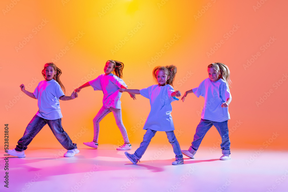 Fototapeta premium Hip-hop dance, street style. Happy children, little active girls in casual style clothes dancing isolated on orange background in purple neon light.