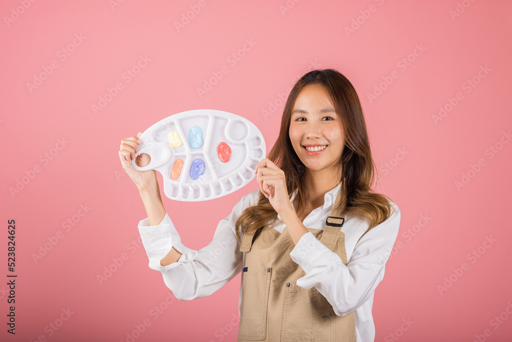 Happy female painting using paintbrush and palette with colors, Asian beautiful young woman artist holding brush and paint palette, studio shot isolated on pink background, Paintings and art equipment Happy female painting using paintbrush and palette with colors, Asian beautiful young woman artist holding brush and paint palette, studio shot isolated on pink background, Paintings and art equipment