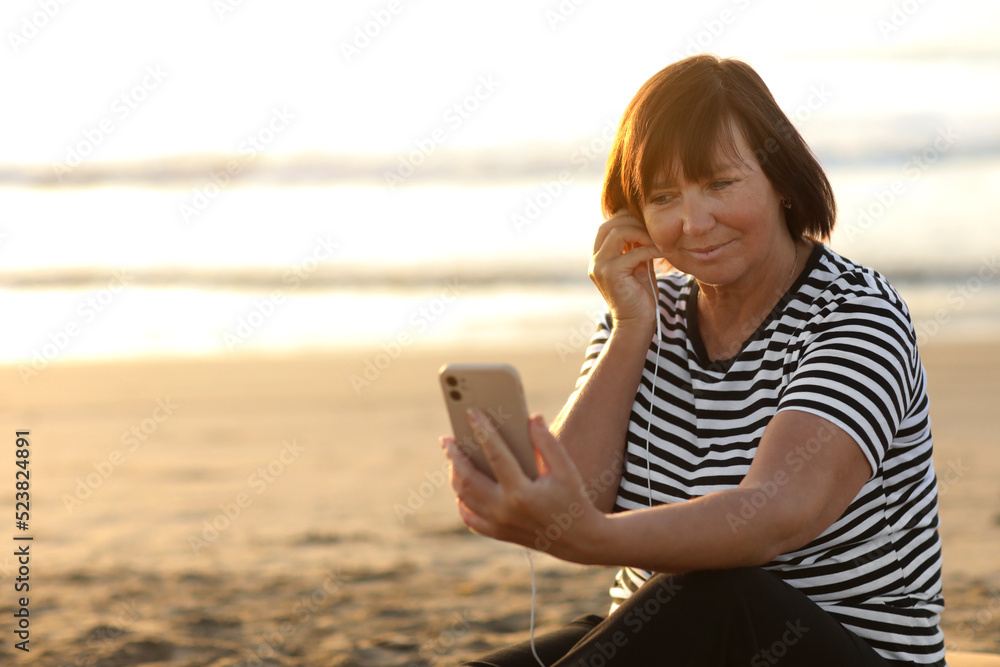 Smiling mature caucasian woman listening music in headphones looking at camera relaxing and resting on sandy beach by the sea at sunrise. Relaxation concept