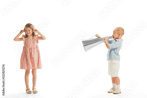 Portrait of little children  stylish boy and girl playing together isolated over white background. Boy screaming in megaphone at girl