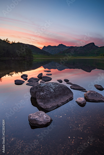 Sunset reflections with rocks in lake and mountains in background at Blea Tarn in the Lake District, UK.