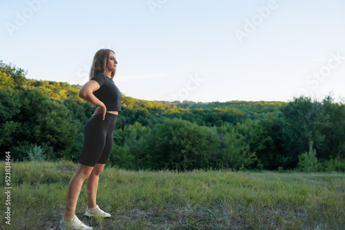a young sportswoman in a black tracksuit keeps her hands on her belt and looks away at the sunset in the park