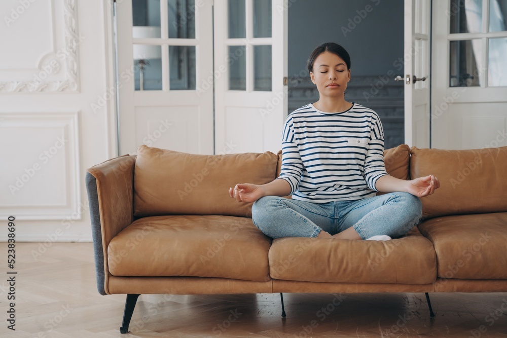 Lady sitting on sofa in lotus asana and meditating in living room. Stress relief and relaxation. Lady sitting on sofa in lotus asana and meditating in living room. Stress relief and relaxation.