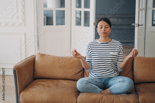 Young woman is practicing yoga at home. Lady is sitting on couch in lotus pose and meditating.