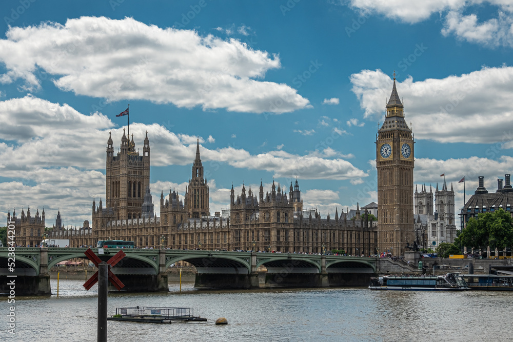 Obraz premium London, UK - July 4, 2022: House of Lords, Westminster Palace, hall and bridge, and Big Ben under blue cloudscape seen over brown water Thames from opposite shore 