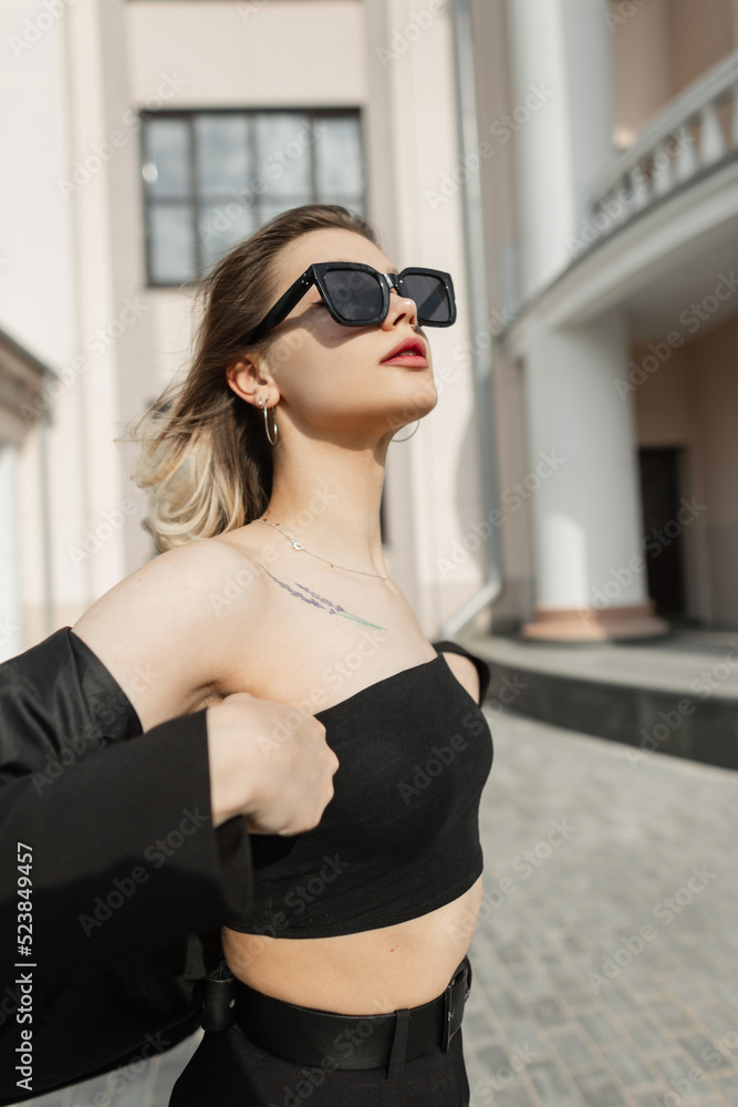 Stylish beautiful young girl with fashion black sunglasses in fashionable clothes with top, blazer and pants walking in the city near a vintage building Stylish beautiful young girl with fashion black sunglasses in fashionable clothes with top, blazer and pants walking in the city near a vintage building