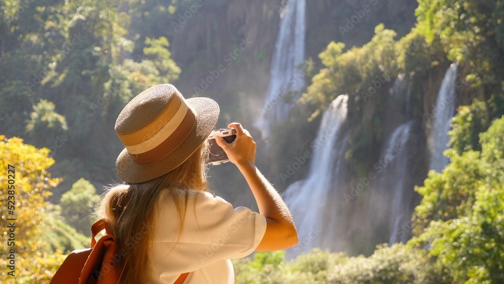 Fototapeta premium Cheerful happy joyful young woman tourist with camera makes photos for social media network or for memories on beautiful tropical waterfall. Concept of active travel, trip in Thailand