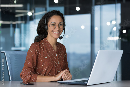 Portrait of young beautiful woman in glasses, looking at camera and smiling Arab woman using headset and laptop for video call, working in modern office, customer support service