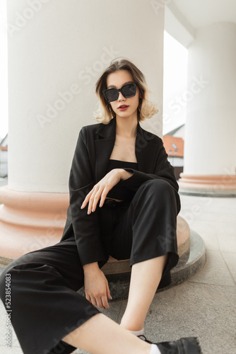 Free Download Fashionable young beautiful business woman model with trendy modern sunglasses in black elegant clothes with blazer, top and pants sits near a vintage column in the city - GFXLoad