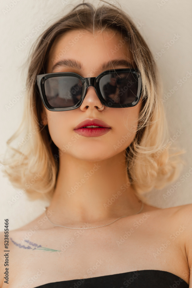 Urban female summer fresh portrait of beautiful young woman model with cool sunglasses in black top stand on a white background on the street Urban female summer fresh portrait of beautiful young woman model with cool sunglasses in black top stand on a white background on the street