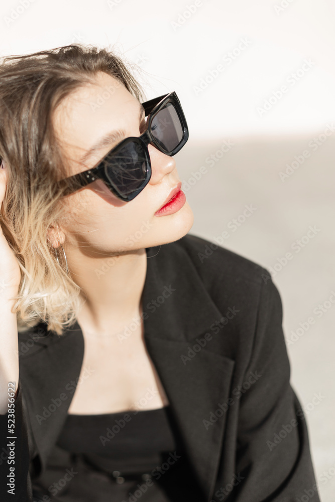 Summer female fresh portrait of beautiful young woman model with cool trendy sunglasses in black elegant clothes sits outdoors at sunlight Summer female fresh portrait of beautiful young woman model with cool trendy sunglasses in black elegant clothes sits outdoors at sunlight