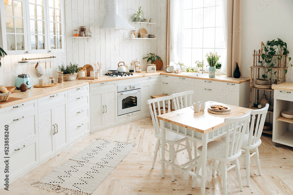 Modern white Scandinavian style kitchen with a wooden worktop filled with kitchen accessories. Stylish kitchen interior with wooden dining table, large windows and potted plants. Empty space