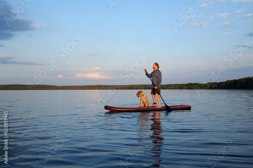 A mature man with a dog is paddling on the lake water on a SUP board. Vacation, tourism, active lifestyle.