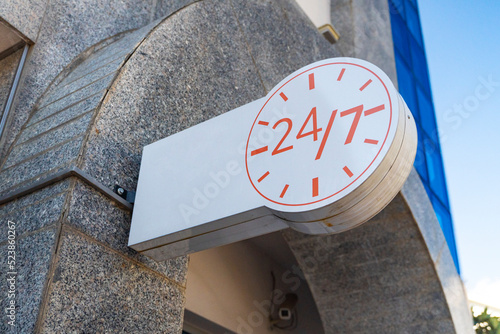 A white banner with the image of a clock signifying the round-the-clock operation of the institution against the background of a blue sky. twenty-four by seven round-the-clock work signage