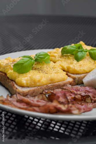 fresh scrambled butter eggs with bacon, pepper and basil on green plate on steel black table 