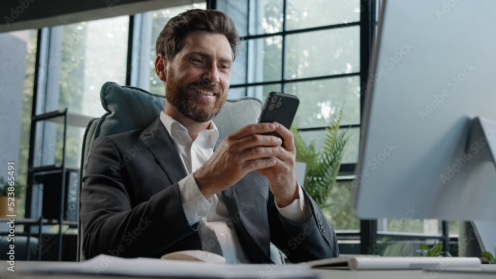 Busy Caucasian bearded man middle-aged 40s businessman uses phone mobile app texting chatting texting message working in office sitting desk with computer remote communicating browsing business chat Busy Caucasian bearded man middle-aged 40s businessman uses phone mobile app texting chatting texting message working in office sitting desk with computer remote communicating browsing business chat
