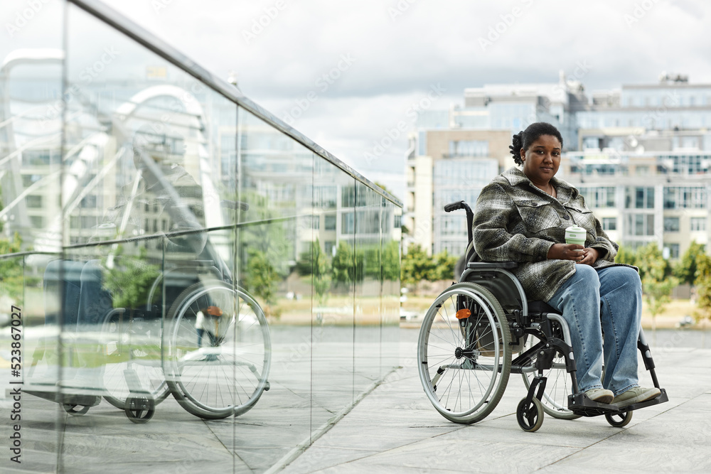Full length portrait of black woman in wheelchair looking at camera outdoors with city skyline in background and glass reflections, copy space
