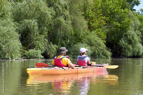 Family couple man and a woman paddle orange kayak near green trees at summer river