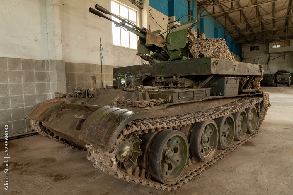 Upgraded T-62 old tank with gun and the Ukrainian flag in the hangar, side view, wide angle shot. An old combat vehicle with an anti-aircraft gun and camouflage net.