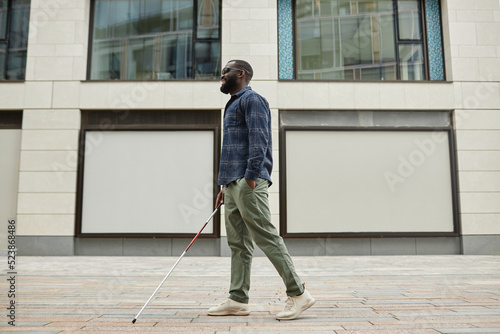 Full length portrait of smiling blind man walking in city and using cane, copy space