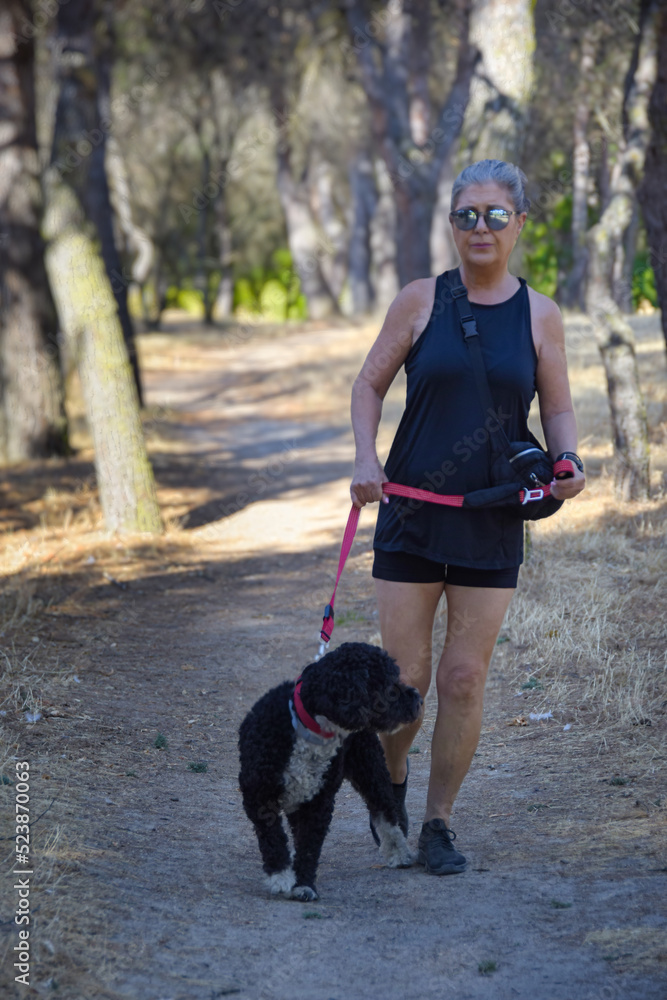 Mujer madura de pelo gris y con gafas de sol paseando por un parque con un perro negro.