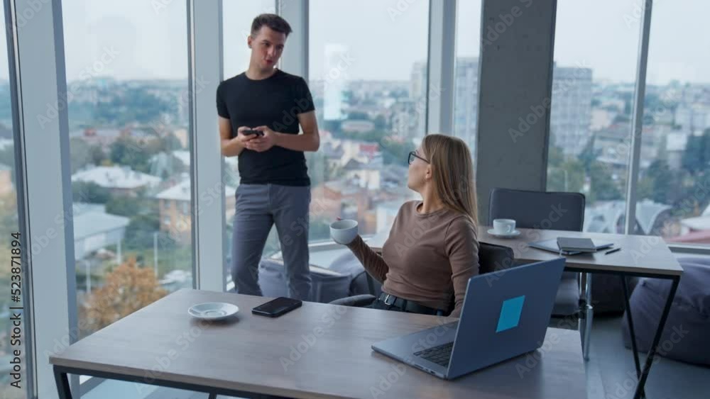 Lunch break in the modern spacious office. Lady is drinking coffee sitting at desk and talking to a male colleague standing at window. Lunch break in the modern spacious office. Lady is drinking coffee sitting at desk and talking to a male colleague standing at window.