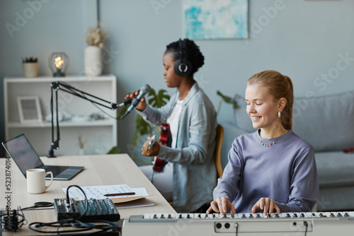 Portrait of smiling young woman playing synthesizer and recording music with friend in home studio, copy space