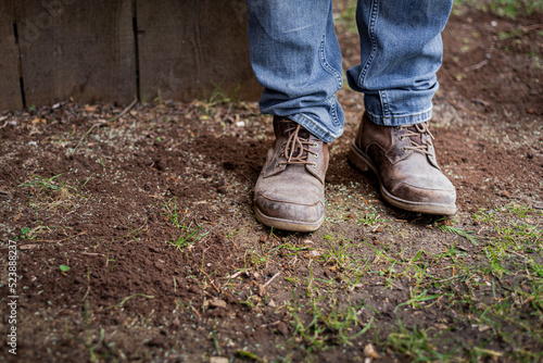 Man's feet and lower legs in blue denim jeans and brown work man's boots standing on patchy garden lawn. 