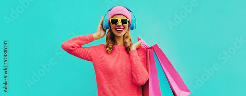 Portrait of stylish happy smiling young woman enjoying listening to music in headphones with colorful shopping bags posing wearing pink knitted sweater, hat on blue background