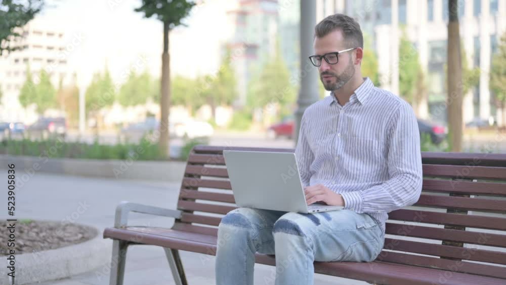 Young Adult Man Talking on Video Call while Sitting Outdoor on Bench Young Adult Man Talking on Video Call while Sitting Outdoor on Bench