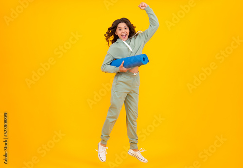 Teen girl dressed in sports uniform. Teenager in a posh stylish sports suit in a hoodie with a hood hold yoga mat. Advertising sportswear tracksuit. Healthy kids lifestyle, sport. © Olena