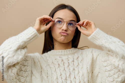 an emotional, funny woman stands in glasses for vision on a beige background holding her glasses with her hands and makes a very funny face, frowning her eyebrows and lowering the corners of her lips