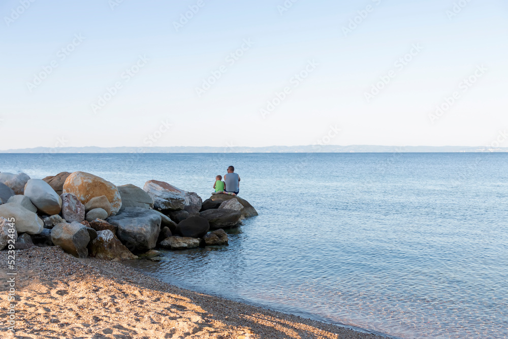 Father and child sitting on the sea coast and enjoying sunny beautiful morning travelling together. Father and lovely son resting in the sunrise Father and child sitting on the sea coast and enjoying sunny beautiful morning travelling together. Father and lovely son resting in the sunrise