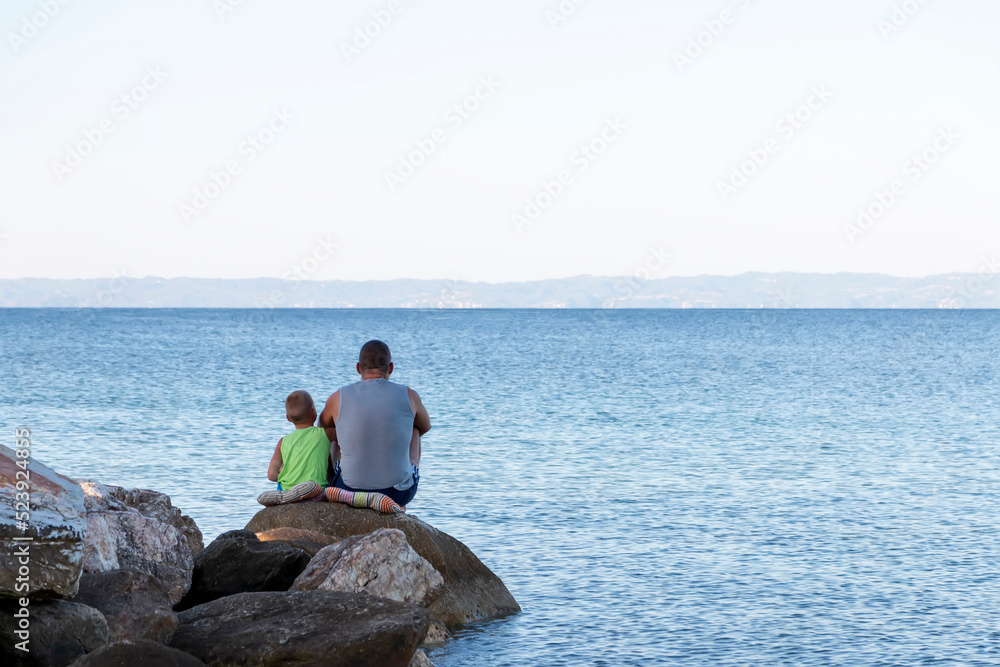 Father and child sitting on the sea coast  and enjoying sunny beautiful morning travelling together. Father and lovely son  resting in the sunrise