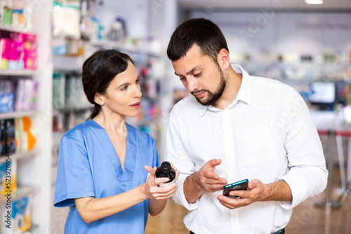 Male shopper shows name of drug on the smartphone screen. Female pharmacist helps to find medicine in pharmacy