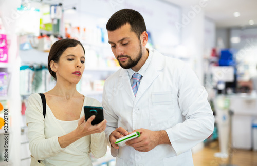 Pharmacist helps to find medicine in a pharmacy by the name on the screen of the buyer smartphone
