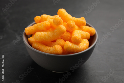 Many tasty cheesy corn puffs in bowl on black table, closeup view