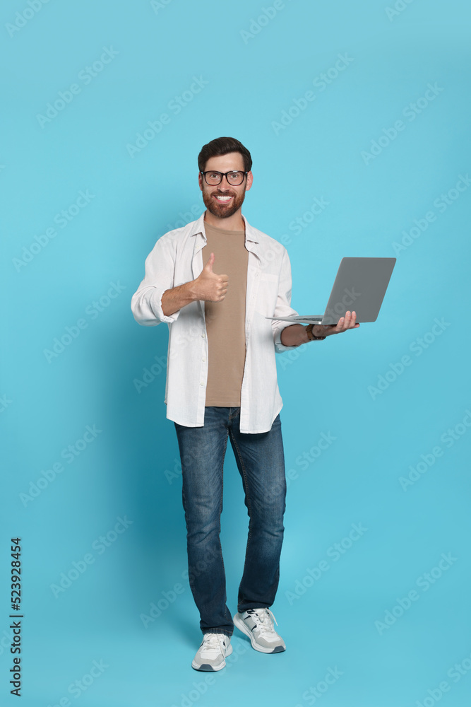 Handsome man with laptop showing thumb up gesture on light blue background