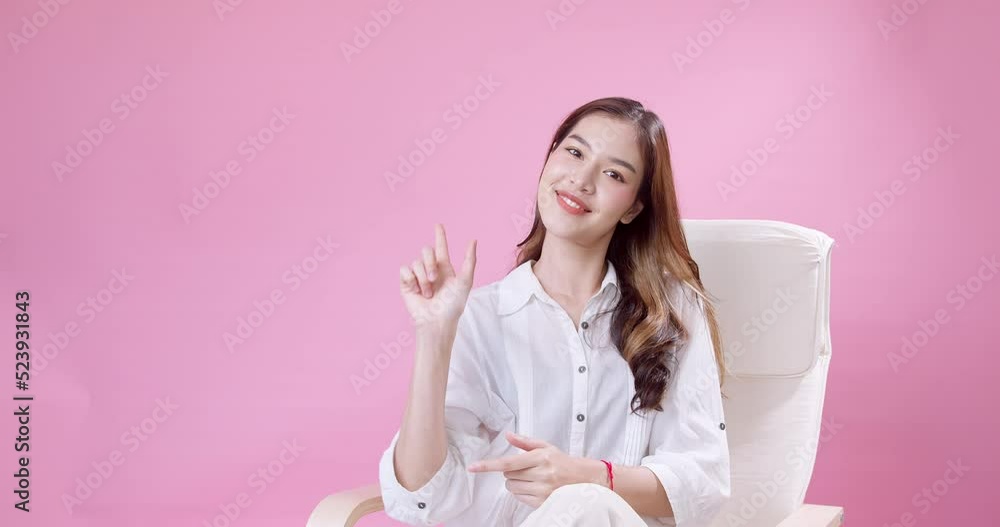 A cheerful, expressive Asian woman in white clothing is sitting on a white chair and pointing with her finger to the side of the object or the empty copy space on isolated pink background. A cheerful, expressive Asian woman in white clothing is sitting on a white chair and pointing with her finger to the side of the object or the empty copy space on isolated pink background.