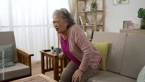 asian senior female having chronic knee pain is sitting back in sofa to massage her joints with a painful look while she is trying to stand up in the living room