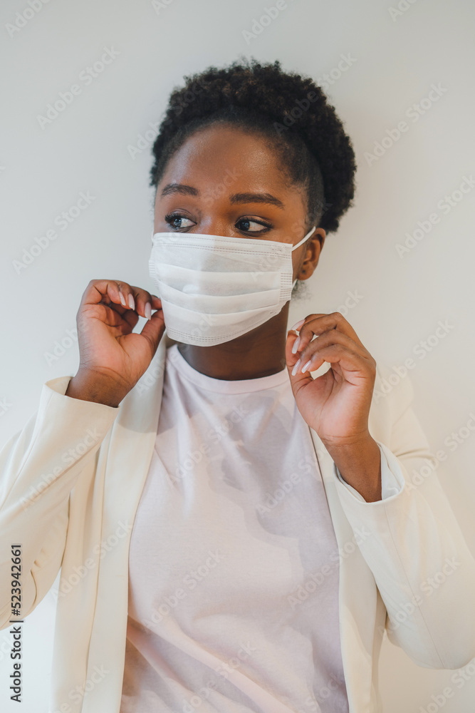 Black female with curly hair putting on medical mask while standing in a room with closed air, and looking away. Isolated over white wall. Respiratory virus Black female with curly hair putting on medical mask while standing in a room with closed air, and looking away. Isolated over white wall. Respiratory virus