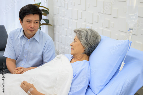 Senior Asian female patient resting on the medical bed in hospital and talking to doctor. 