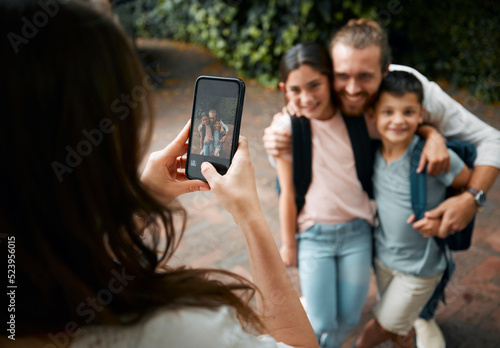 Mother taking a family photo on her phone of her happy little children and their father smiling before school. Boy and girl siblings with their dad smile for a picture. Mom taking photos of kids