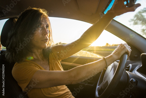 Hispanic lady driving a car on the road while adjusting the rear view mirror at sunset time. Copy space. © ideasRojas 